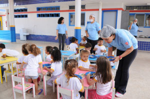 Crianças sentadas em mesa infantil recebem merenda, oferecida por funcionários da creche