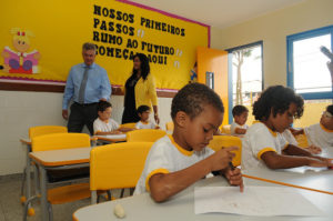 Crianças pequenas estudam em cadeiras e mesas infantis, quanto governador visita a sala de aula