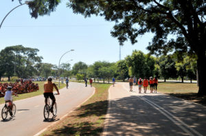 A pista de caminhada nova paralela à pista antiga de ciclismo com pessoas caminhando e ciclista pedalando, céu azul, dia ensolarado, árvores verdes. 