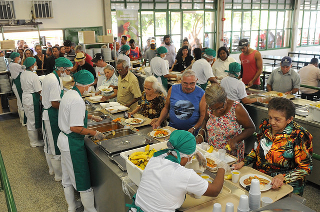 Pessoas na fila do restaurante comunitário sendo servidas. Na foto, há vários idosos.