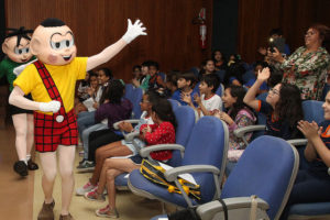 Os personagens Cascão e Cebolinha acenam para crianças em auditório