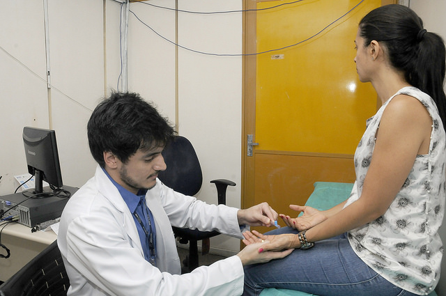 na foto, um médico realiza um exame na mão de uma paciente sentada em uma maca