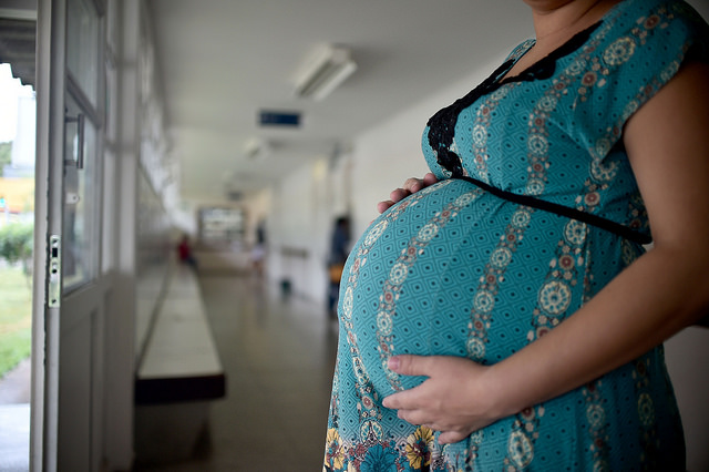 na foto, uma mulher grávida de vestido azul mostra a barriga