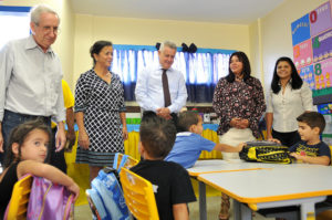 Governador, secretário e professoras em pé conversam com alunos sentados em carteiras infantis dentro de sala em creche.