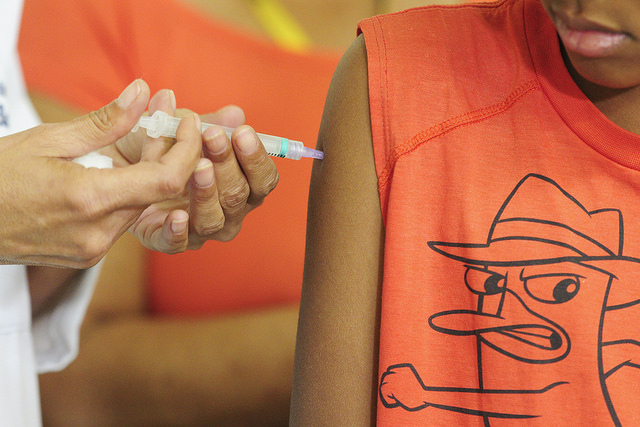 na foto, uma criança vestida com uma camiseta laranja recebe uma vacina no braço