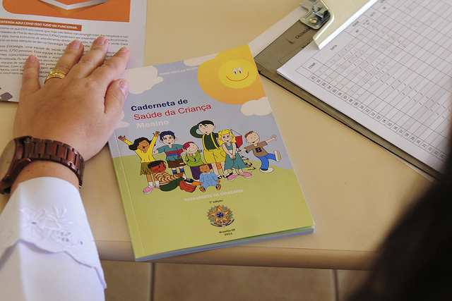 foto de um braço de um médico ao lado de uma caderneta com desenhos de criança e escrito "caderneta de saúde da criança"