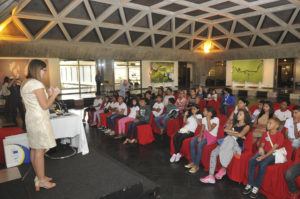 crianças sentadas escutam professora falando em auditório