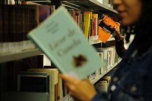 Na imagem aparece uma menina segurando um livro aberto em uma biblioteca. A imagem está desfocada. 