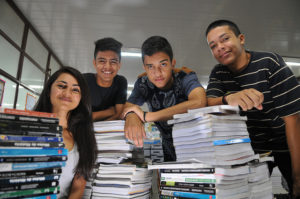 Foto de quatro estudantes aprovados na UNB em frente a uma pilha de livros