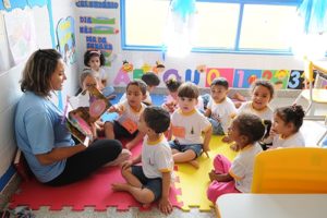 Professora e crianças sentados no chão colorido dentro de sala na creche