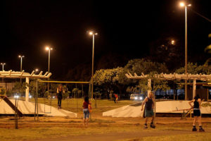 À noite, em parque infantil, uma criança brinca no balanço e alguns adultos fazem caminhada